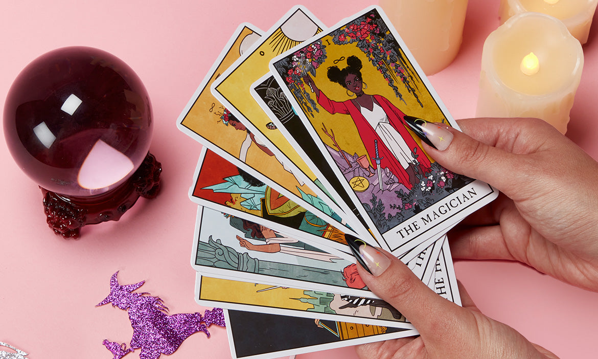 Press-on nails displayed alongside tarot cards, a crystal ball, glowing candles, and glittery bat decorations on a pink background.