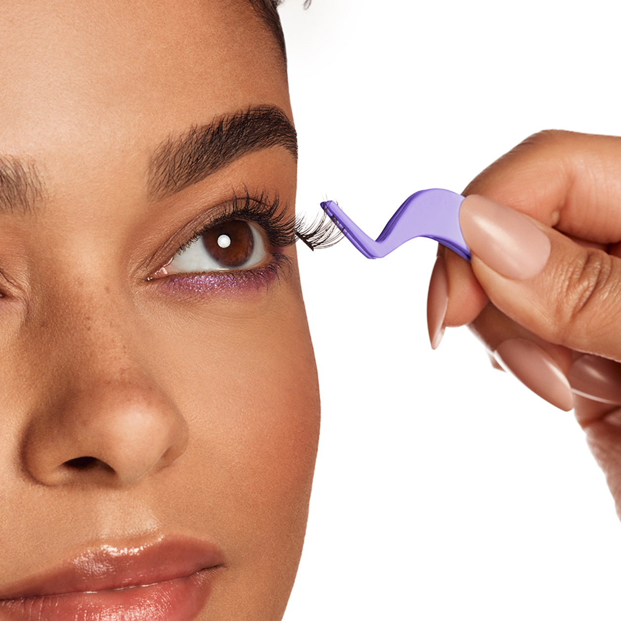 Close-up of a model applying false eyelashes using a purple applicator, with soft glam makeup and a subtle purple eyeliner accent.