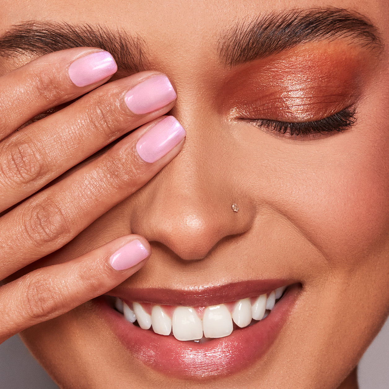 Smiling model with shimmery copper eyeshadow, glossy lips, and pastel pink nails, covering one eye with her hand.