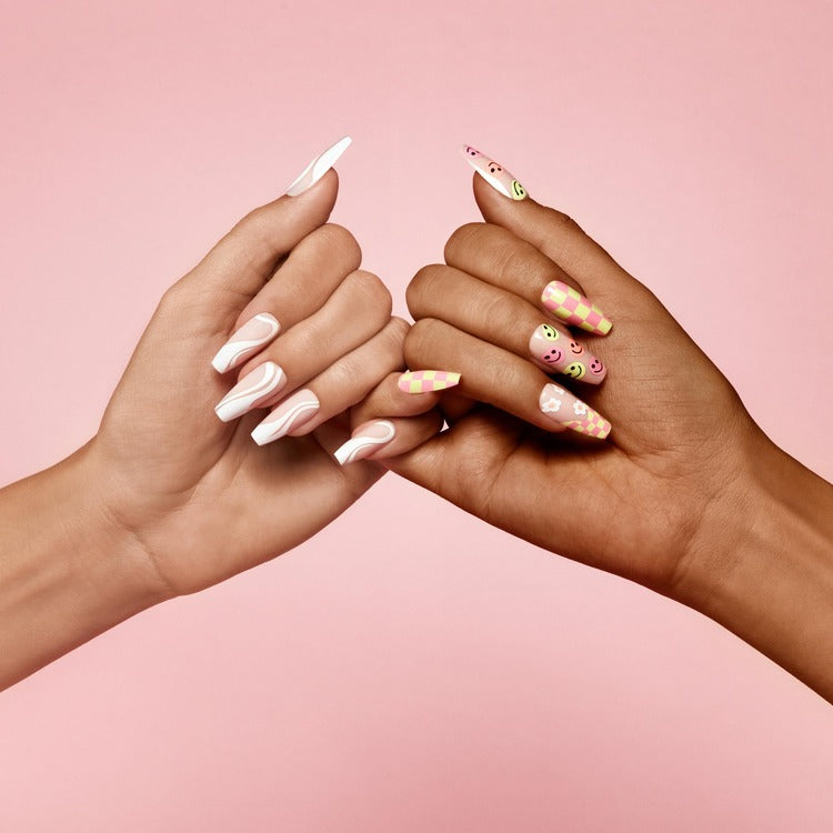 Two hands with trendy press-on nails—one with white swirls, the other with pink checkered and smiley designs, against a pink background.