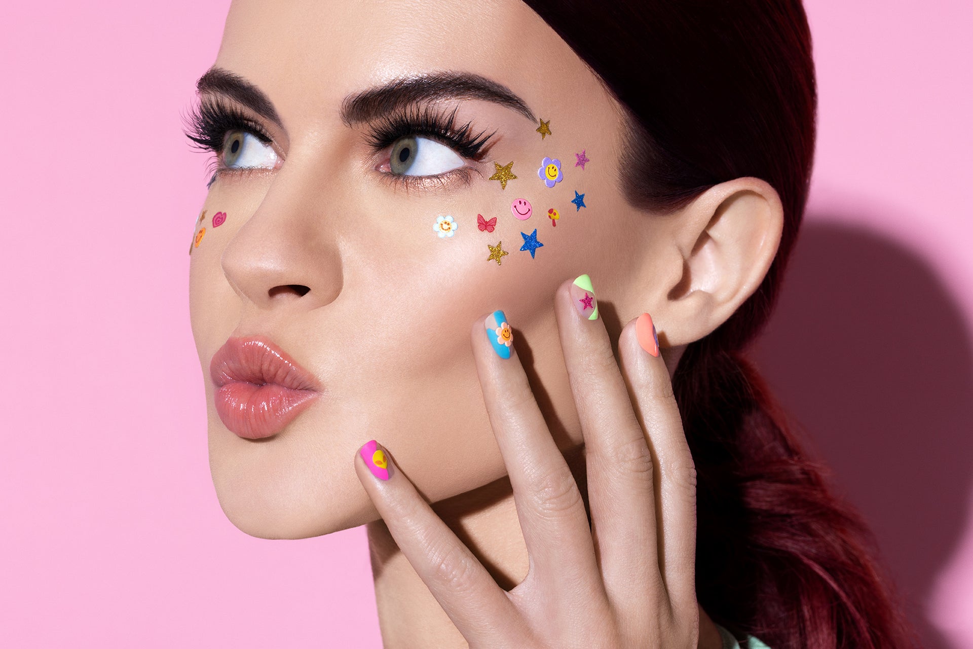 Model with playful face stickers, colorful press-on nails, glossy lips, and bold lashes, posing against a pink background.